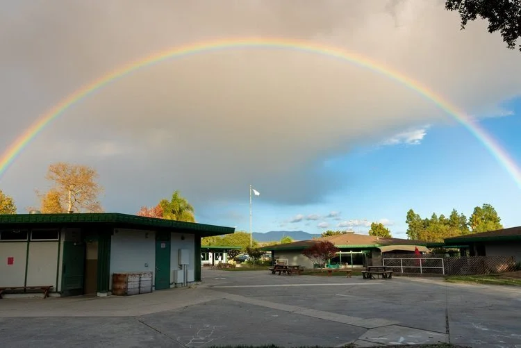 A rainbow arching across the sky over a school courtyard with single-story buildings, picnic tables, trees, and a flagpole.