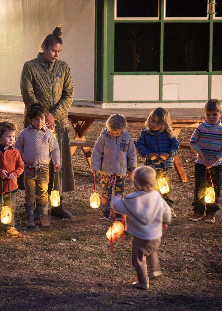 Children and an adult holding lanterns at night outside near a building with a large window.