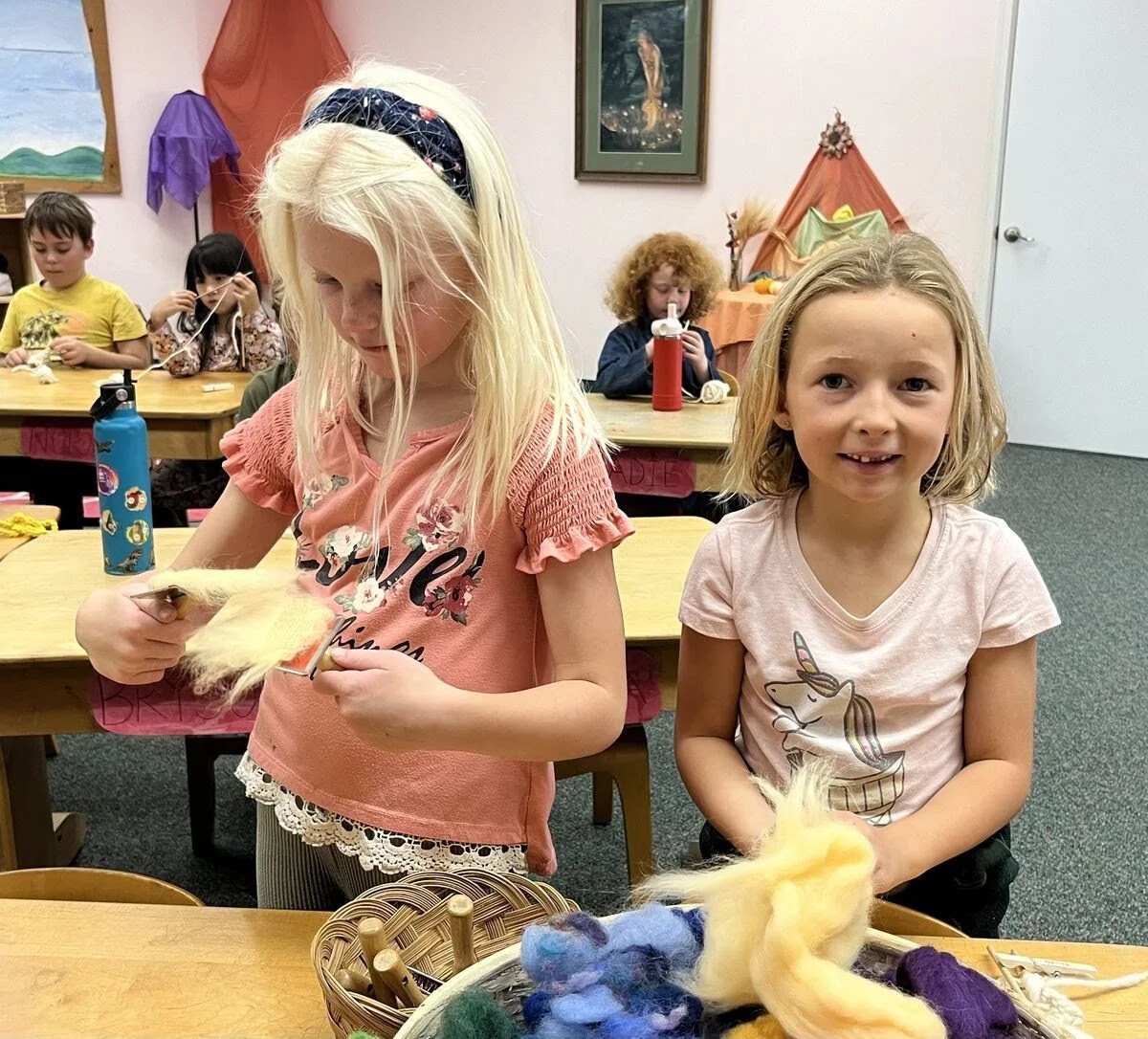 Two young girls in a classroom engaged in a craft activity with wool, surrounded by other children. One girl with blonde hair and wearing a pink shirt is looking down at her project, while the other girl with curly hair and a unicorn shirt is facing 