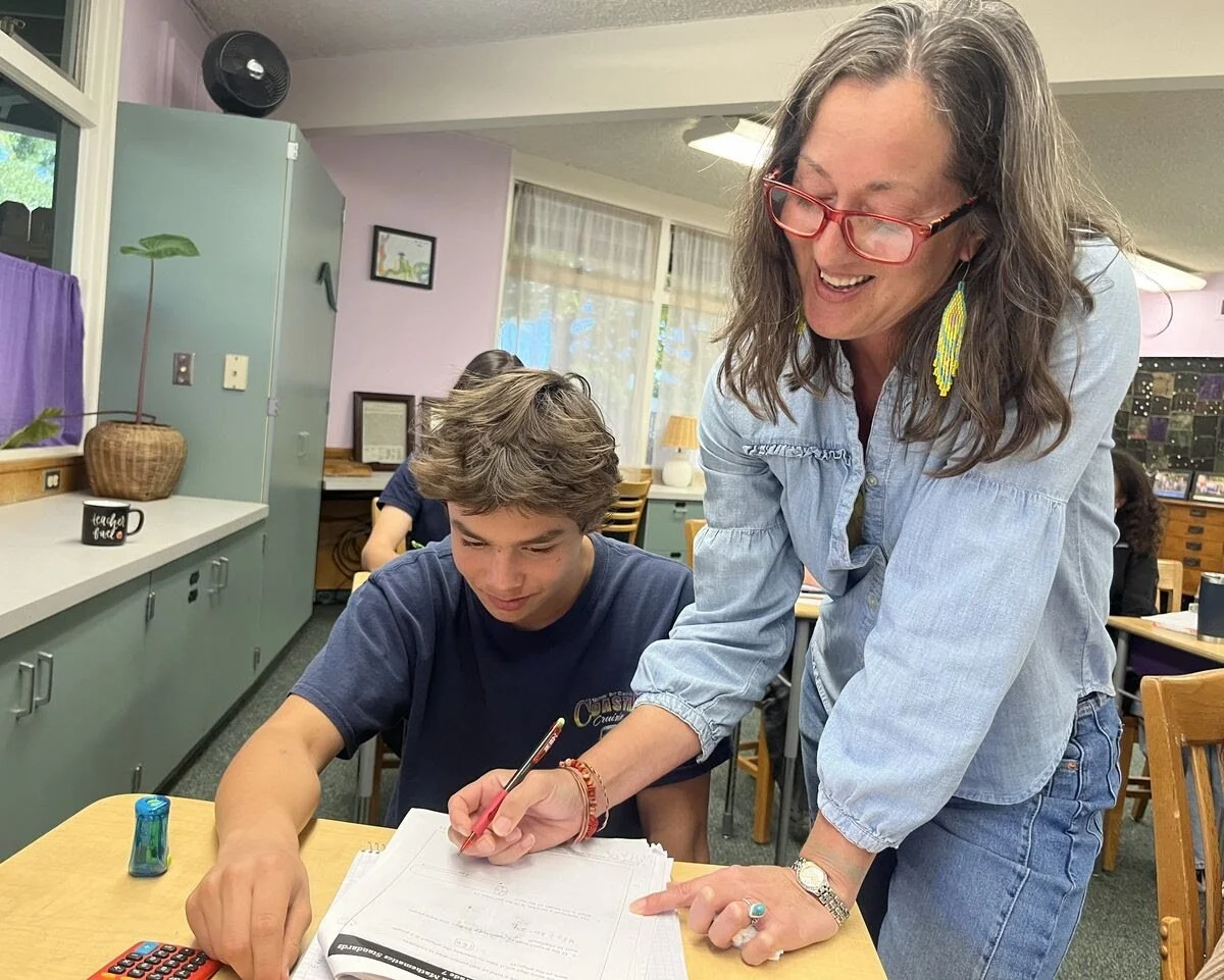 A woman helping a young boy with schoolwork at a school desk in a classroom.