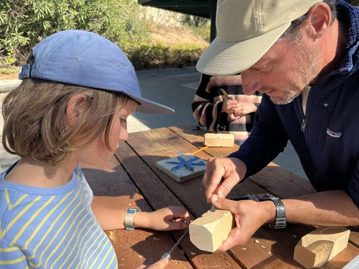A man and a girl at a wooden table outdoors, working together on a woodworking project with wood and tools.