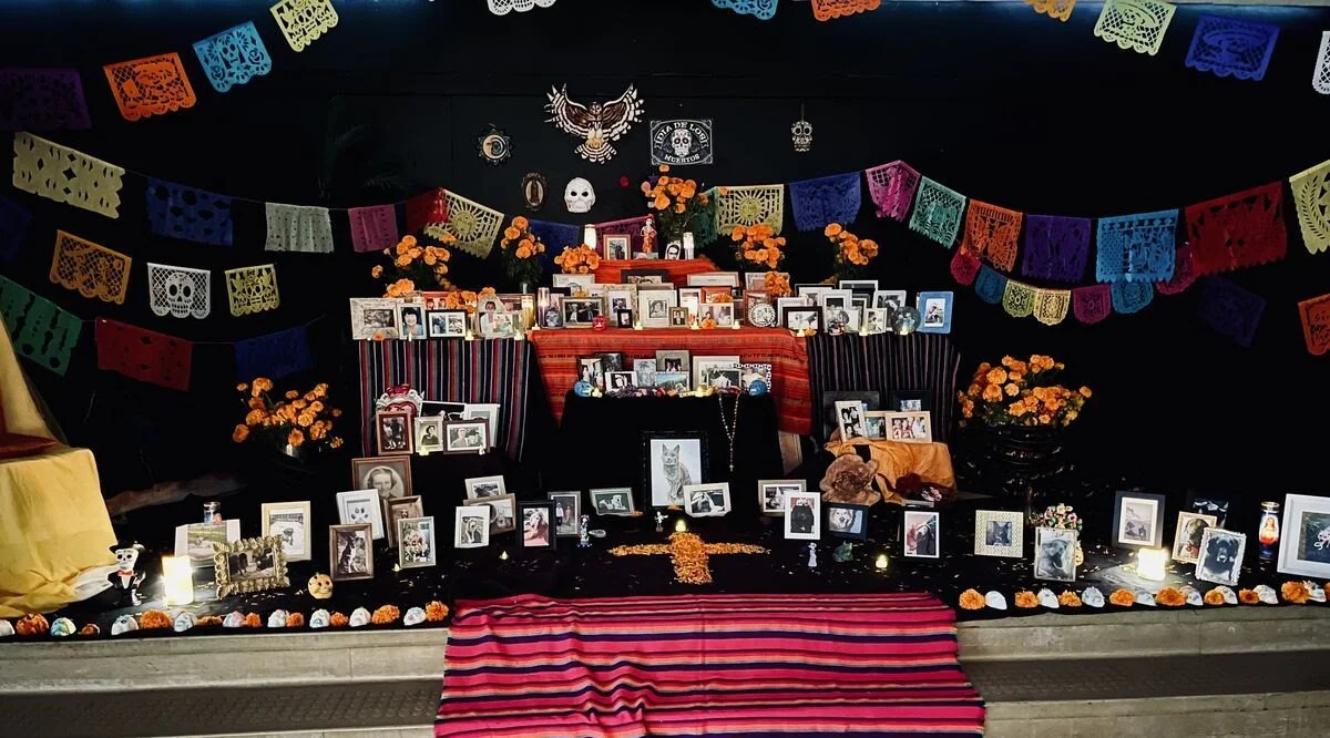Memorial ofr Dia de los Muertos featuring framed photographs of loved ones, colorful papel picado banners, marigold flowers, candles, and decorative items on a black background.