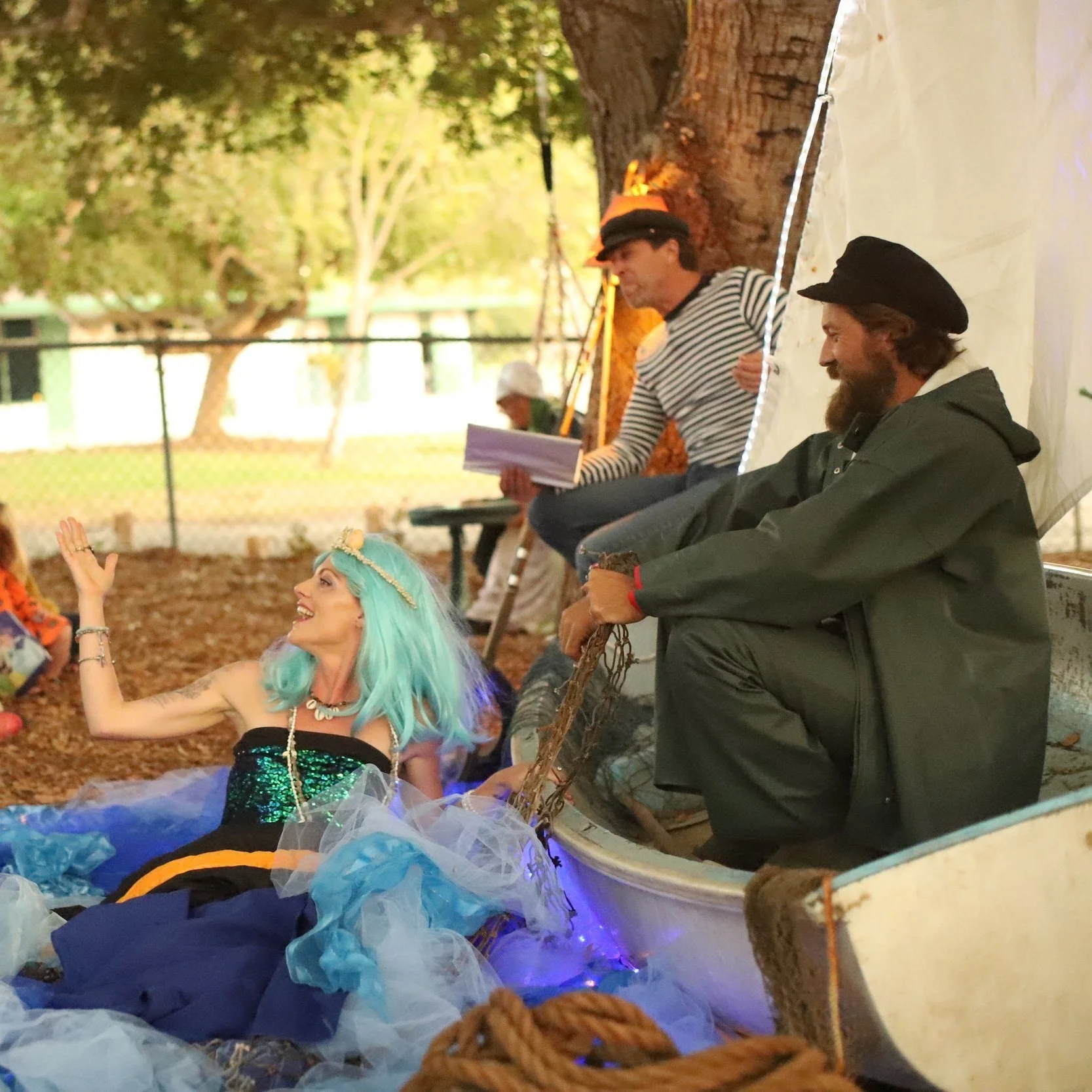 Group of people engaging in a theatrical outdoor performance beneath a tree. A woman with blue hair dressed in a colorful, fantasy-style costume sits in a boat, smiling and waving. Two men, one in a striped shirt and the other with a beard in dark cl