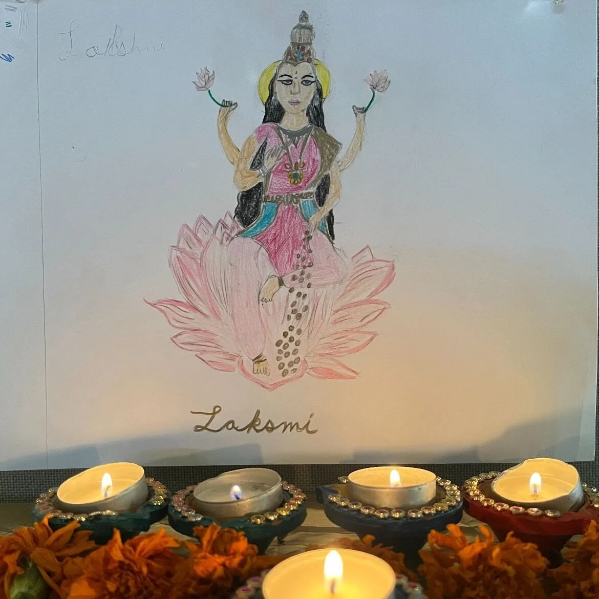 A hand-drawn picture of the Hindu goddess Lakshmi, sitting on a pink lotus flower with four hands, two holding lotus flowers, and wearing traditional attire with jewelry, with her name written below. In front of the drawing are decorated oil lamps an