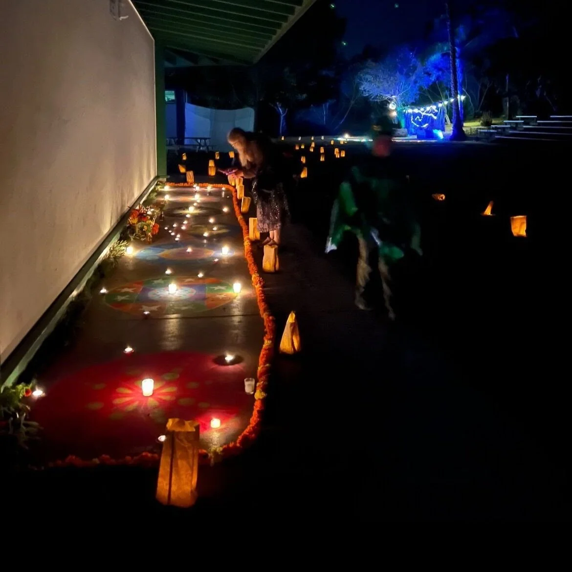 People lighting candles and placing flowers on a decorated outdoor altar at night, with illuminated candles and decorative rugs, celebrating a cultural or religious event.