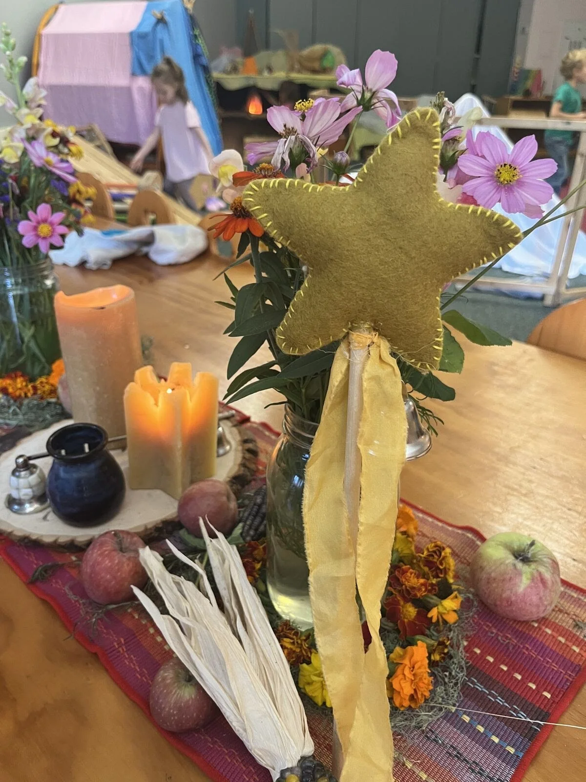 A table decorated with flowers, apples, candles, and a star-shaped felt decoration on a stick. In the background, children are playing and activities are visible.