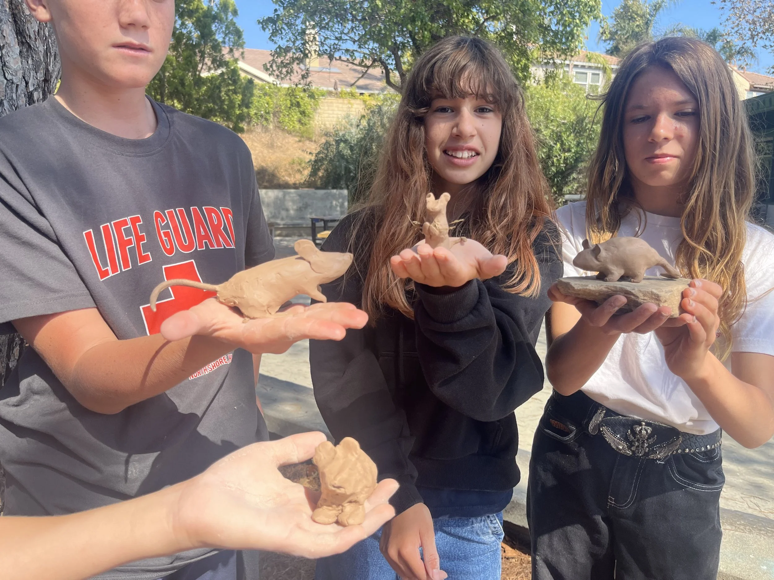 Three children outdoors holding small sculptures of animals, including a rat, a bear, and a mouse, made of clay.
