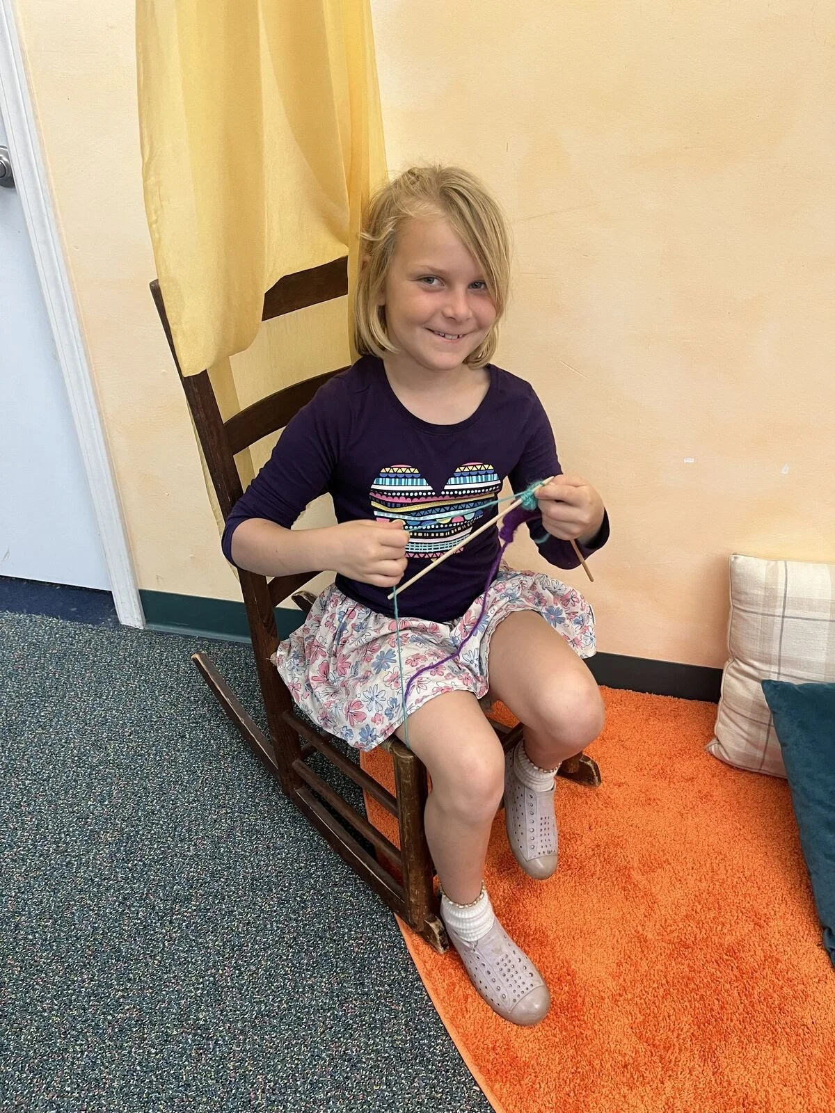 A young girl with blonde hair, smiling, sitting on a wooden rocking chair while knitting with light blue and purple yarn.