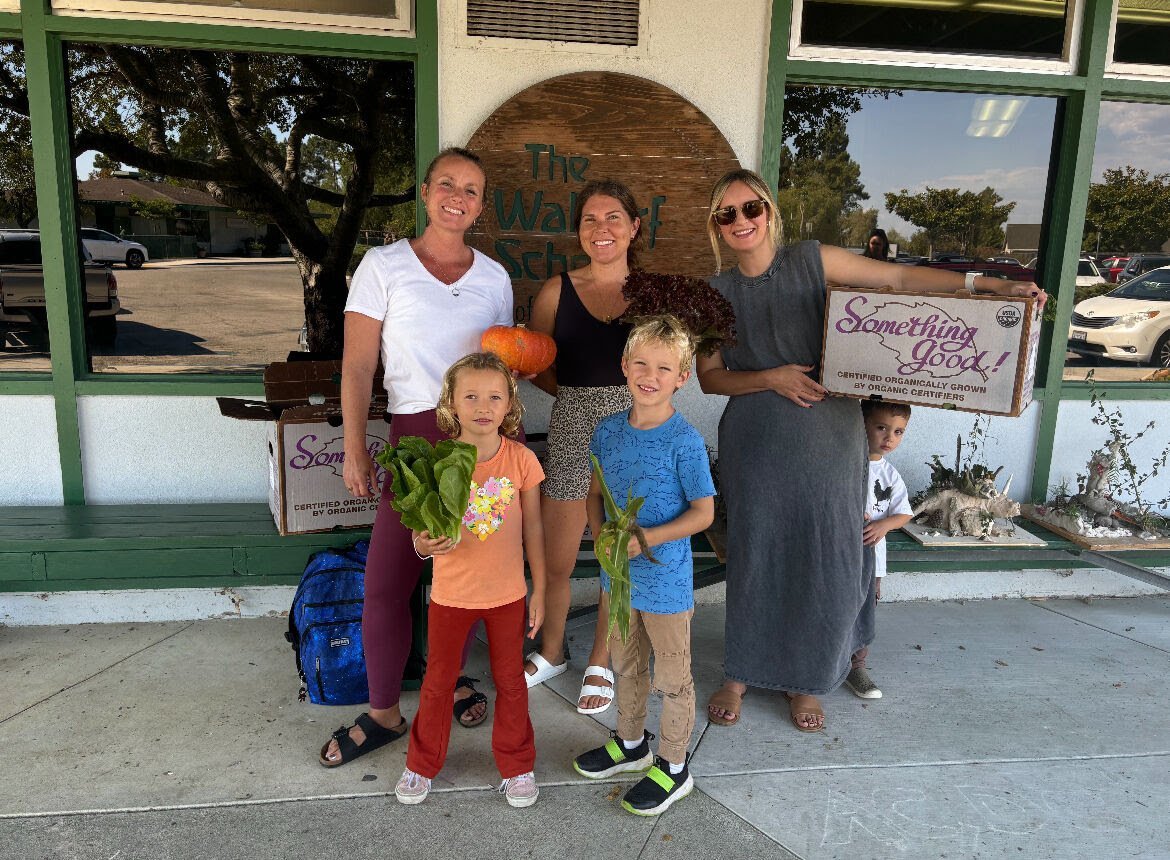 Group of five people, including three women and two children, standing outside a building with a sign that reads "The Waldorf School." The women are holding boxes of organic produce, and the children are holding leafy greens and a pumpkin. One woman 