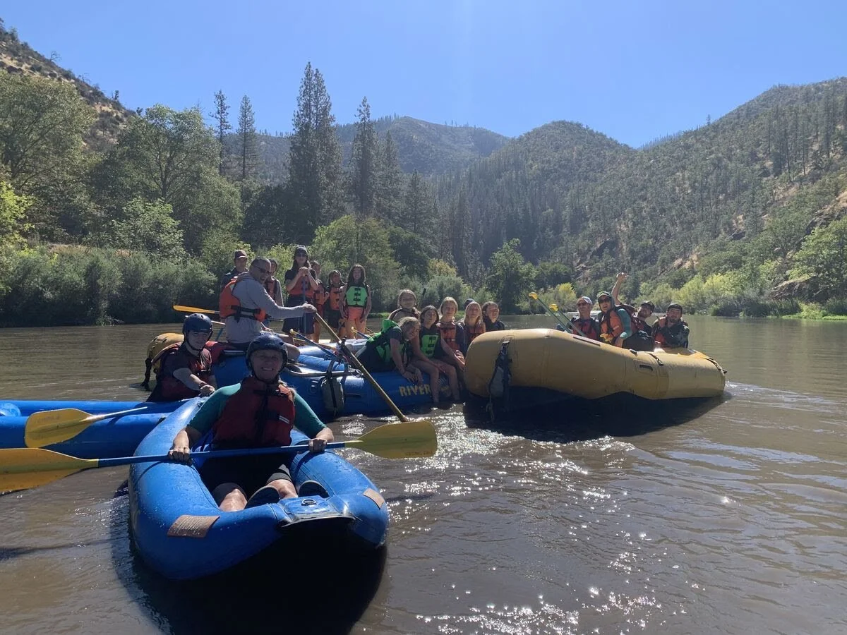 Group of people on inflatable rafts floating on a river surrounded by trees and mountains on a sunny day.
