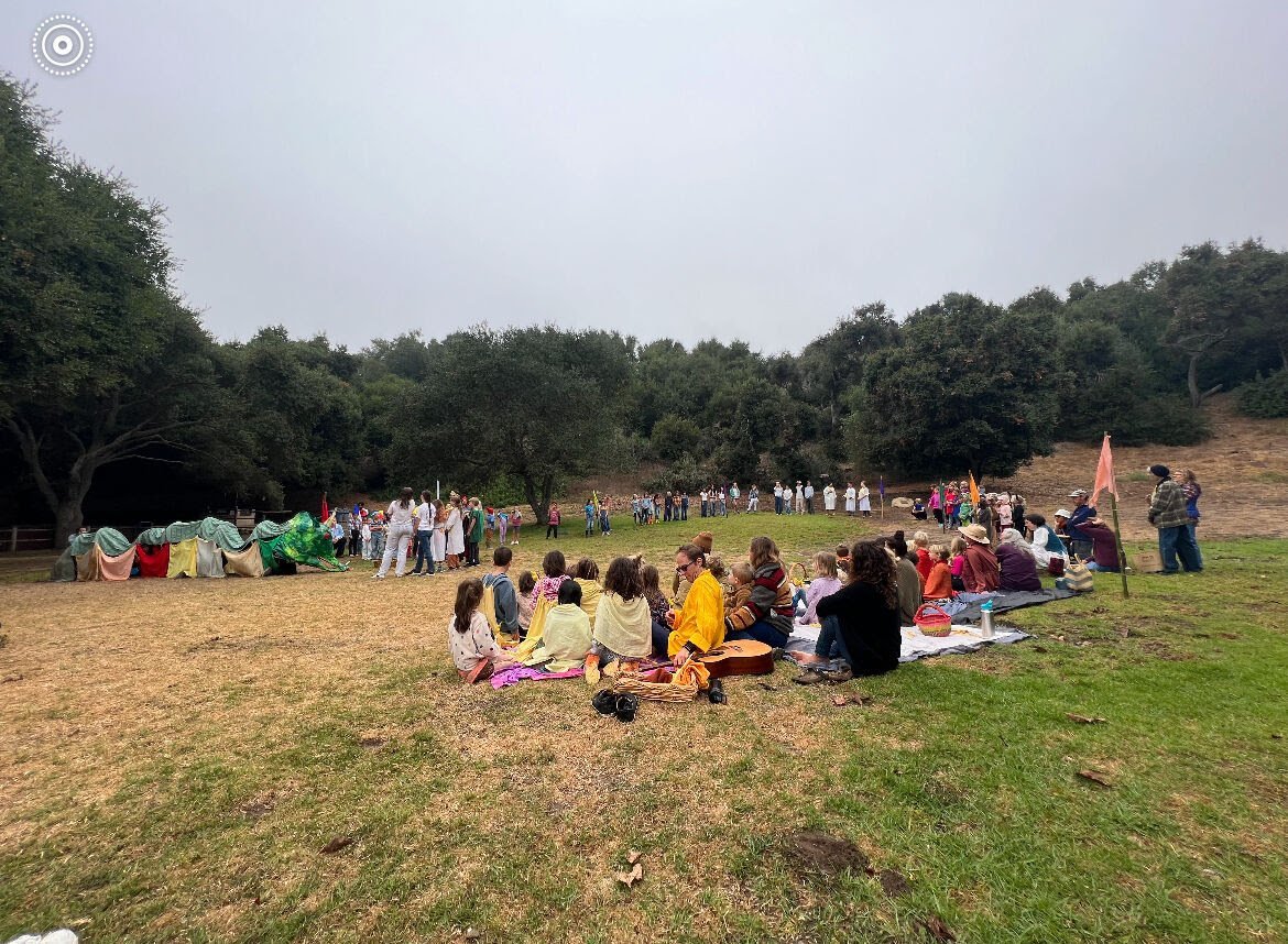 A large outdoor gathering with people seated on the grass and others standing in a semi-circle, likely watching a performance or event, set in a park or natural area with trees and cloudy sky.