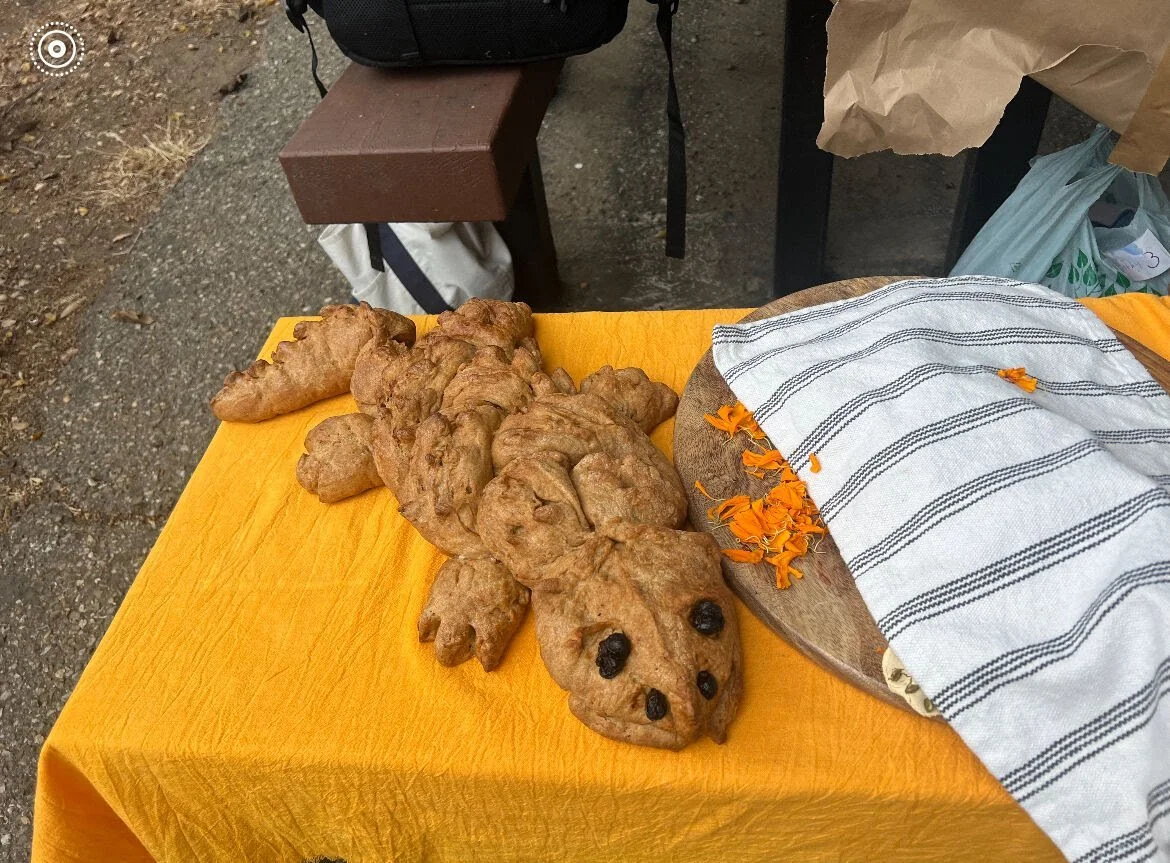 An altar with a gingerbread lizard, a round bread loaf with orange flower petals, and a striped cloth on top, set on a yellow tablecloth outdoors.
