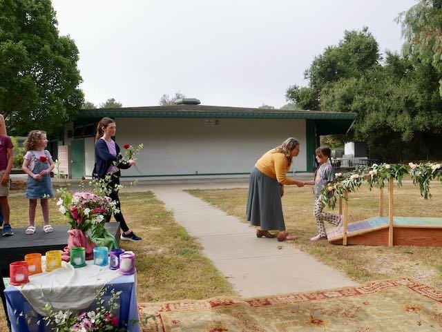 A woman presenting an award or certificate to a young girl at an outdoor ceremony, with children and adults watching.
