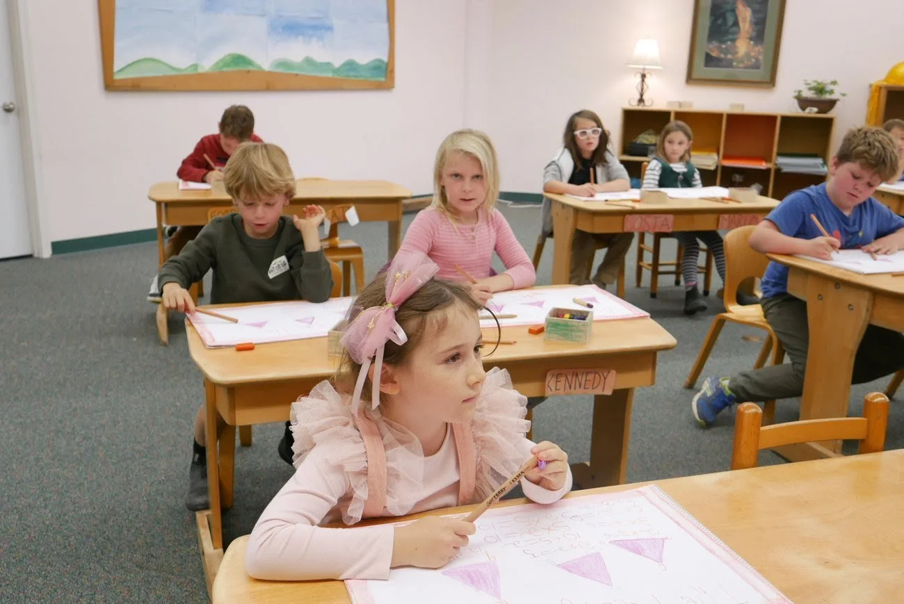Children sitting at desks in a classroom, working on drawing sheets with pink triangle shapes, some children are focused while others are looking around, with colorful pictures on the wall behind them.