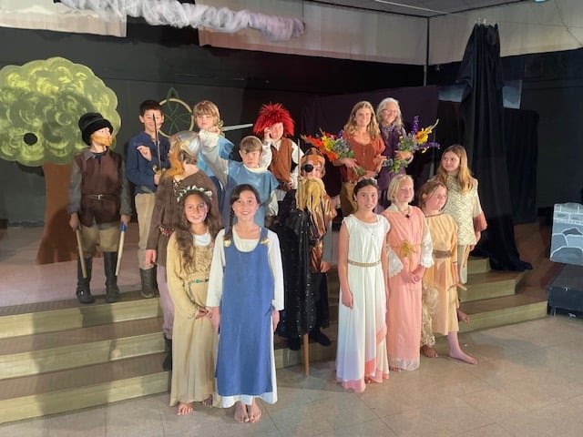 Children performing in a school play, dressed in costumes representing characters from a fairy tale or story, with some dressed as princes, princesses, and other storybook characters, standing on stage with set decorations including a painted tree an