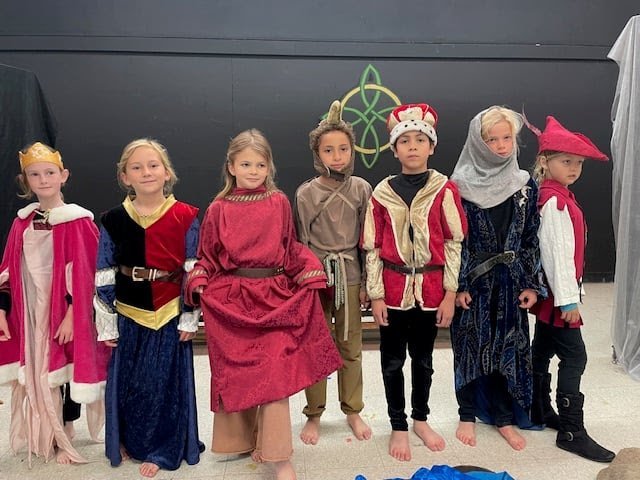 Seven children dressed in medieval or fantasy costumes, standing in a line indoors against a dark wall with a green Celtic knot design, portraying royalty, knights, and peasants.