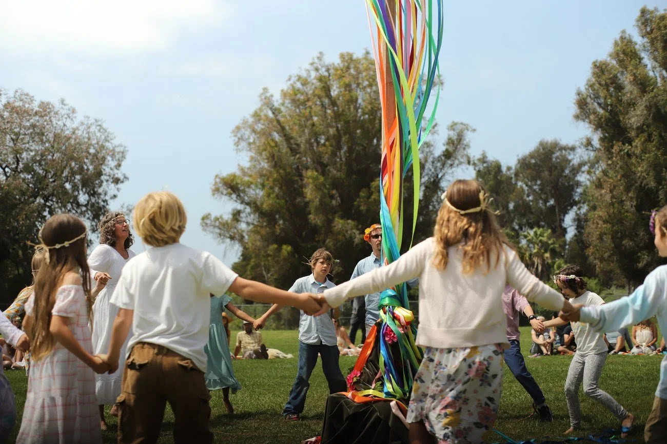 Children and adults holding hands in a circle, dancing around a large colorful ribbon pole outdoors on a sunny day with trees in the background.