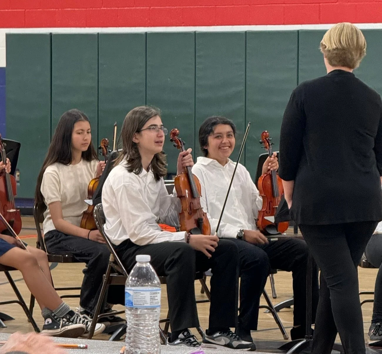 Young orchestra musicians sitting with violins in a concert hall, listening to a conductor, with a water bottle on the floor in front.