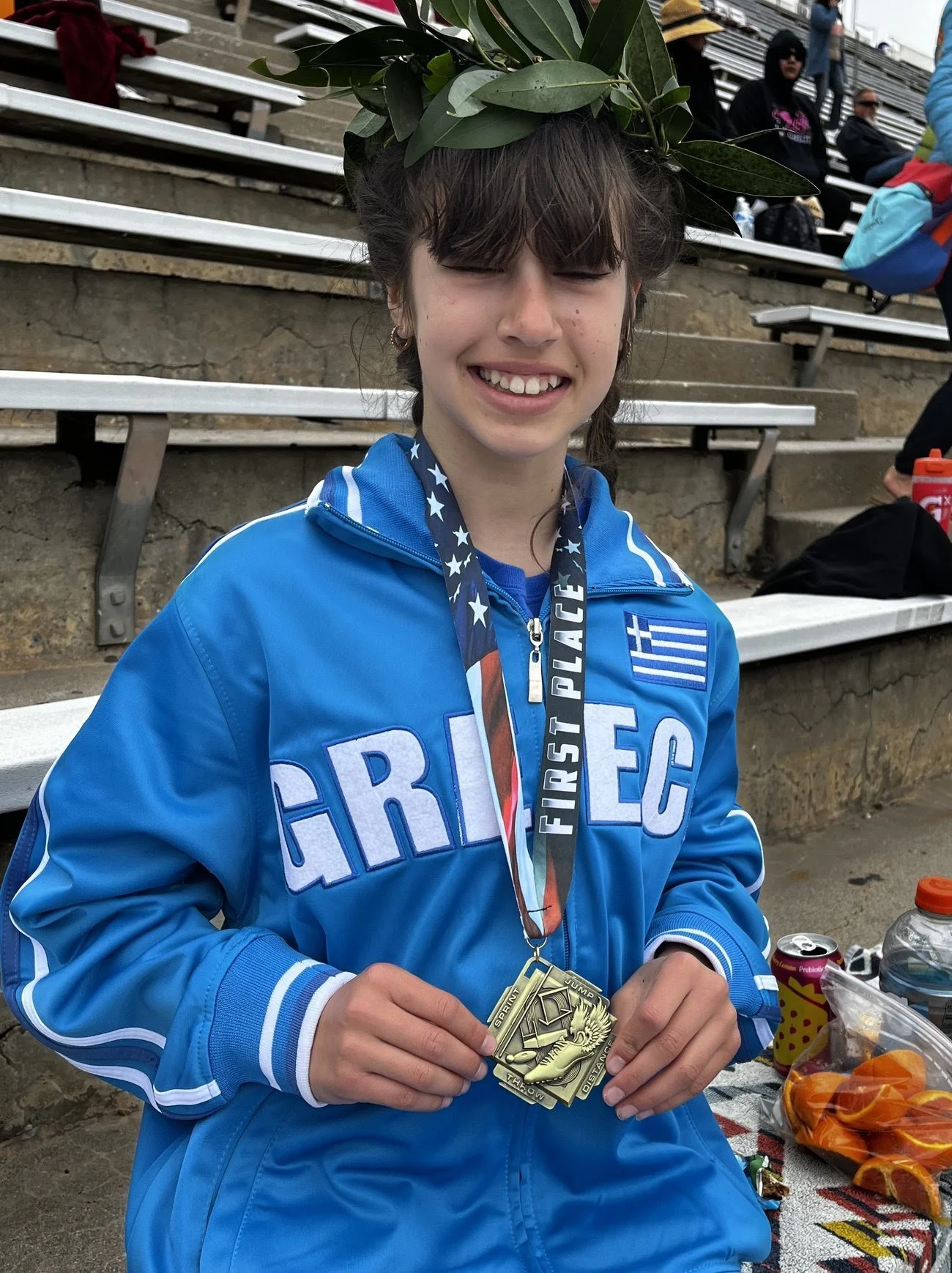 A young girl wearing a blue jacket with Greek flag and text 'GREECE' on it, holding medals, and wearing a safety medal around her neck with a laurel crown on her head, sitting on metal bleachers at an outdoor event.