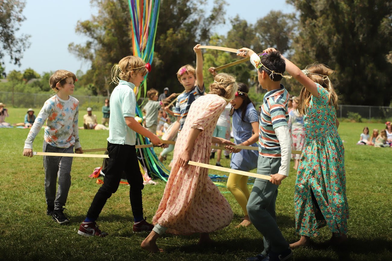 Children playing a game of tug-of-war outdoors on a grassy field with trees and spectators in the background.