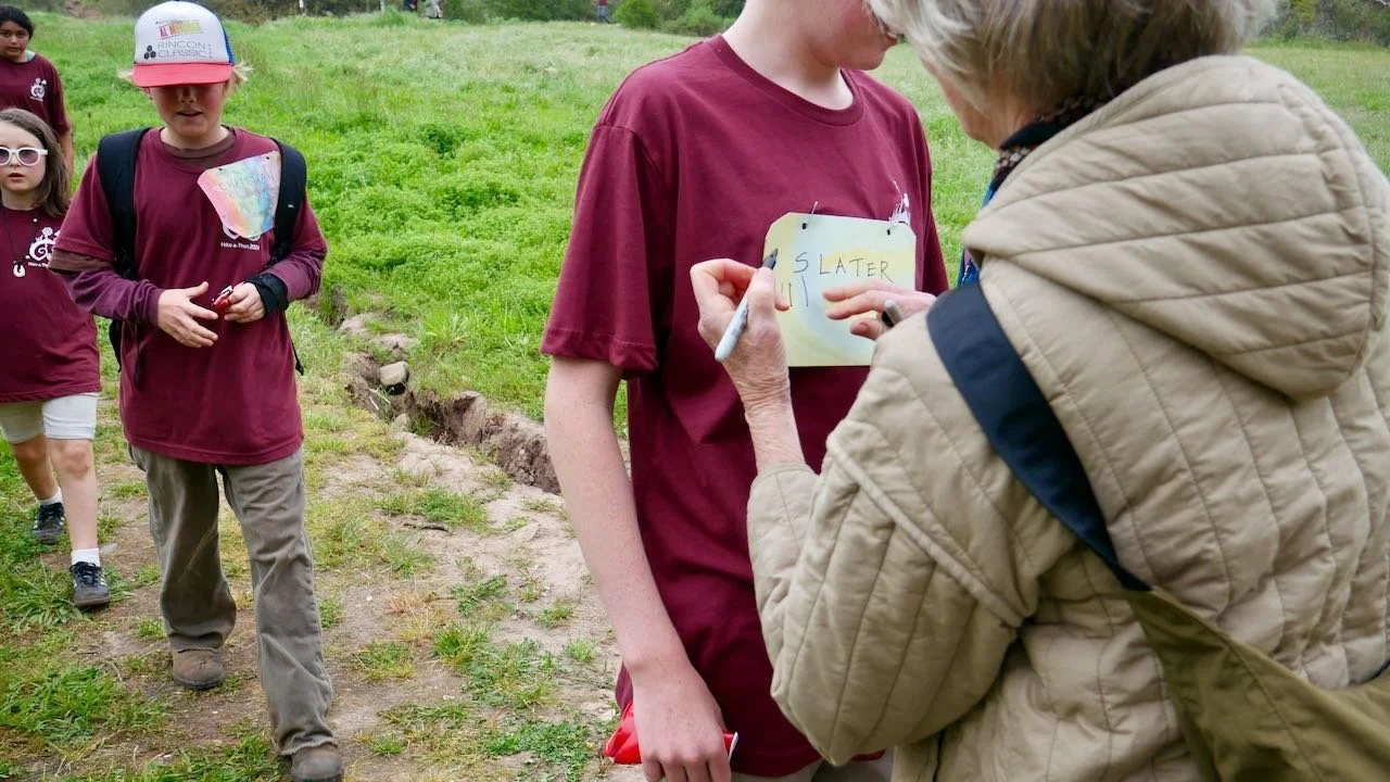 A group of children outside on a grassy area, with one child having a sign pinned to their shirt that says 'S LATER'. An older woman is writing something on the sign with a marker, while other children observe.
