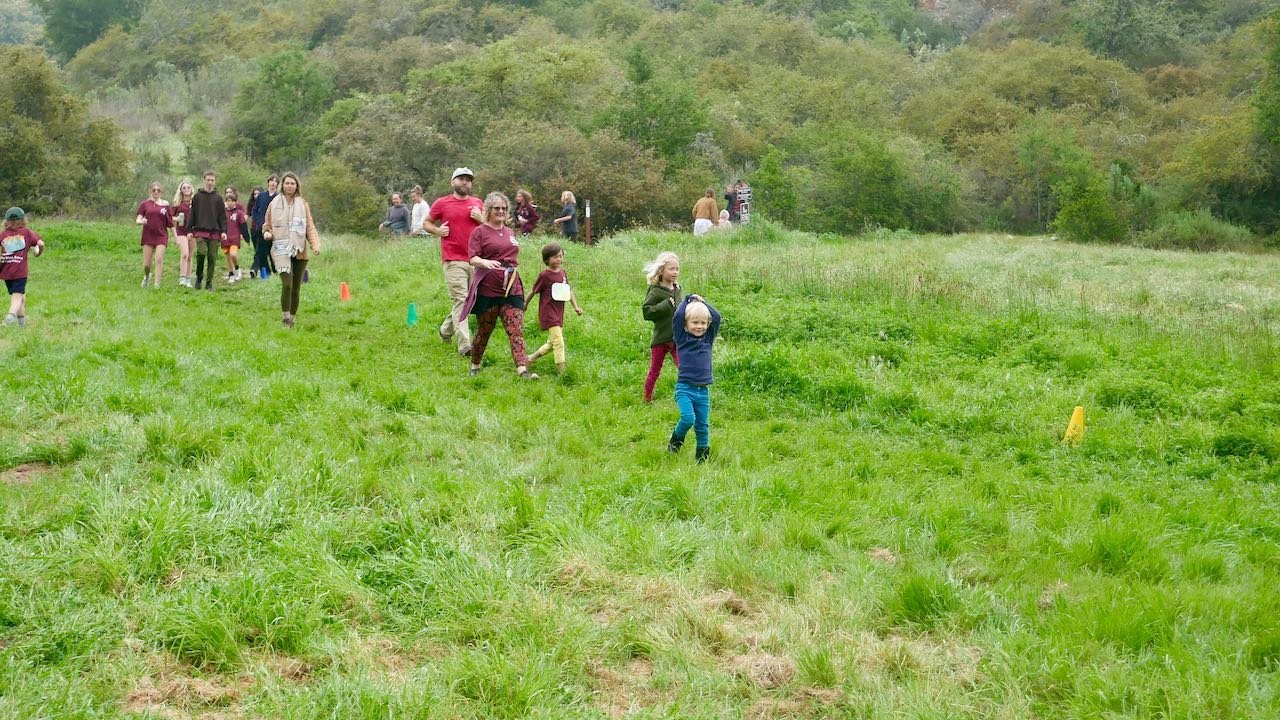 Group of children and adults walking in a grassy field, some children are wearing burgundy shirts, with trees and hills in the background.