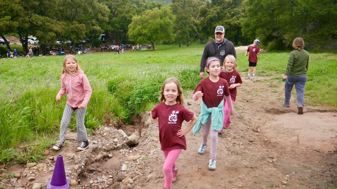 Children and an adult walking outdoors in a park, some children wearing maroon shirts with a logo, others in casual clothing, with trees and other people in the background.