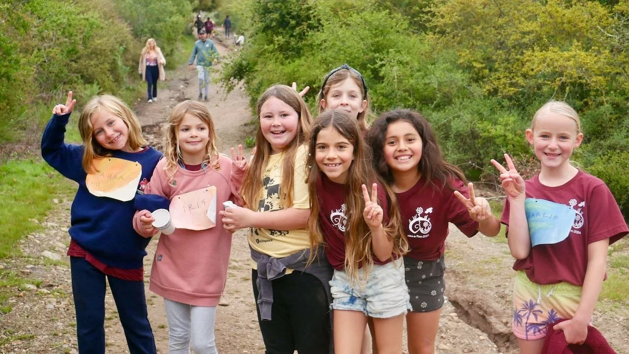Seven young girls, smiling and making peace signs, walking on a trail surrounded by green trees.