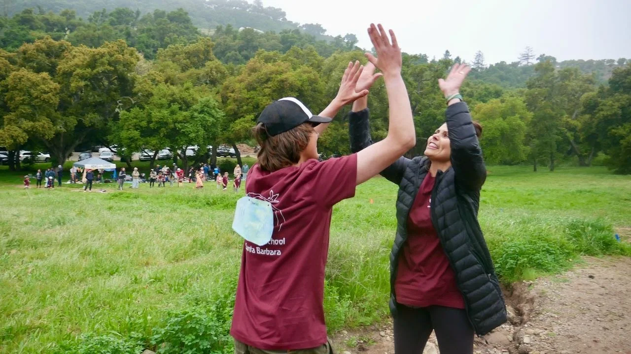 Two women high-fiving in a grassy park with a crowd and trees in the background.