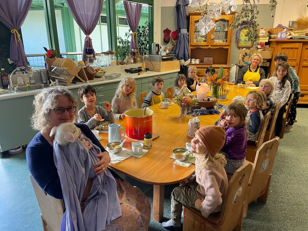 A group of children and two adults gathered around a large wooden table in a cozy kitchen, enjoying a meal. The table is decorated with flowers and various dishes. The room has large windows, purple curtains, and kitchen cabinets and shelves filled w