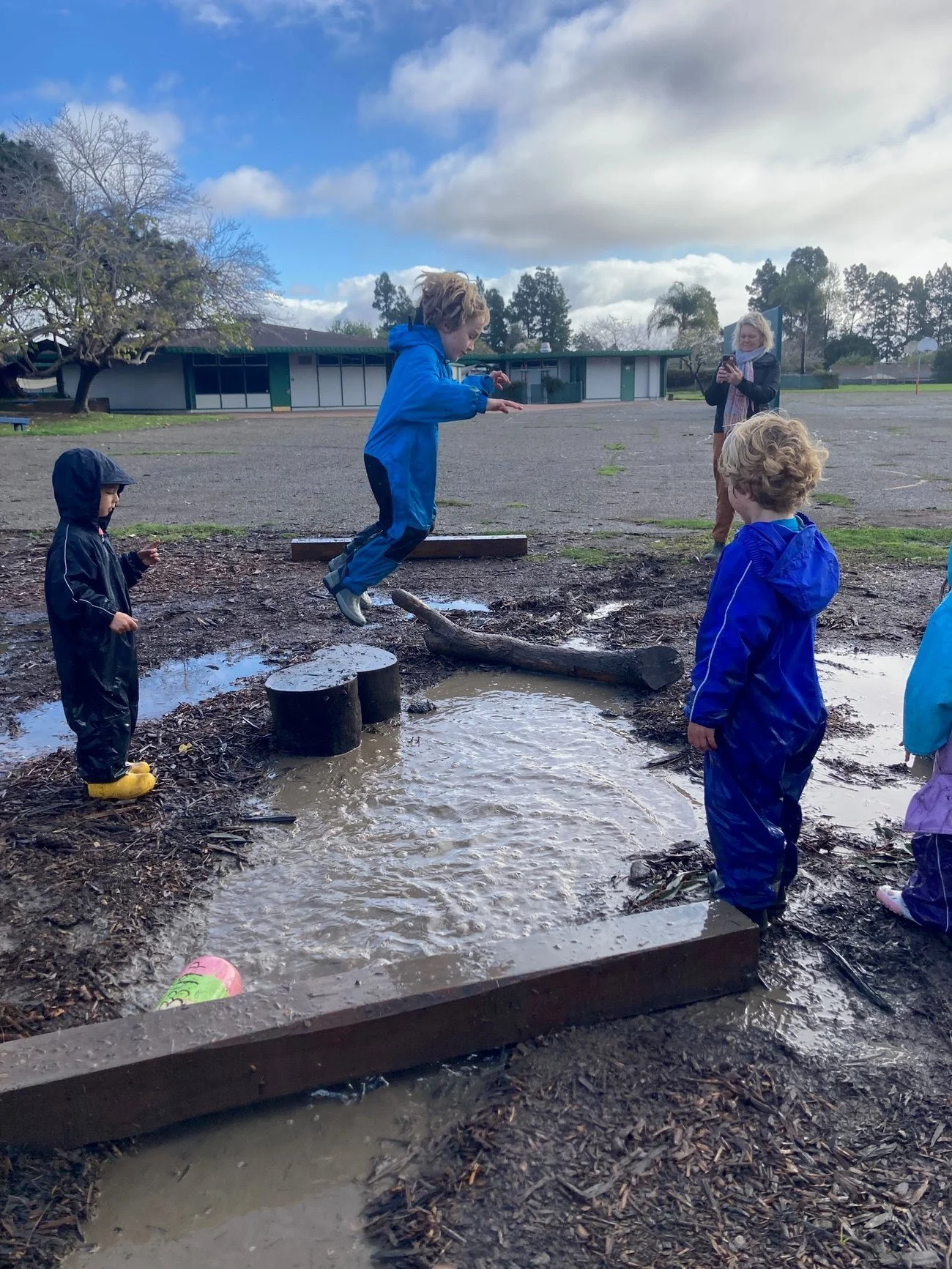 Children playing on a muddy playground, one jumping over a small puddle, others watching, with a woman taking photos in the background, cloudy sky overhead.