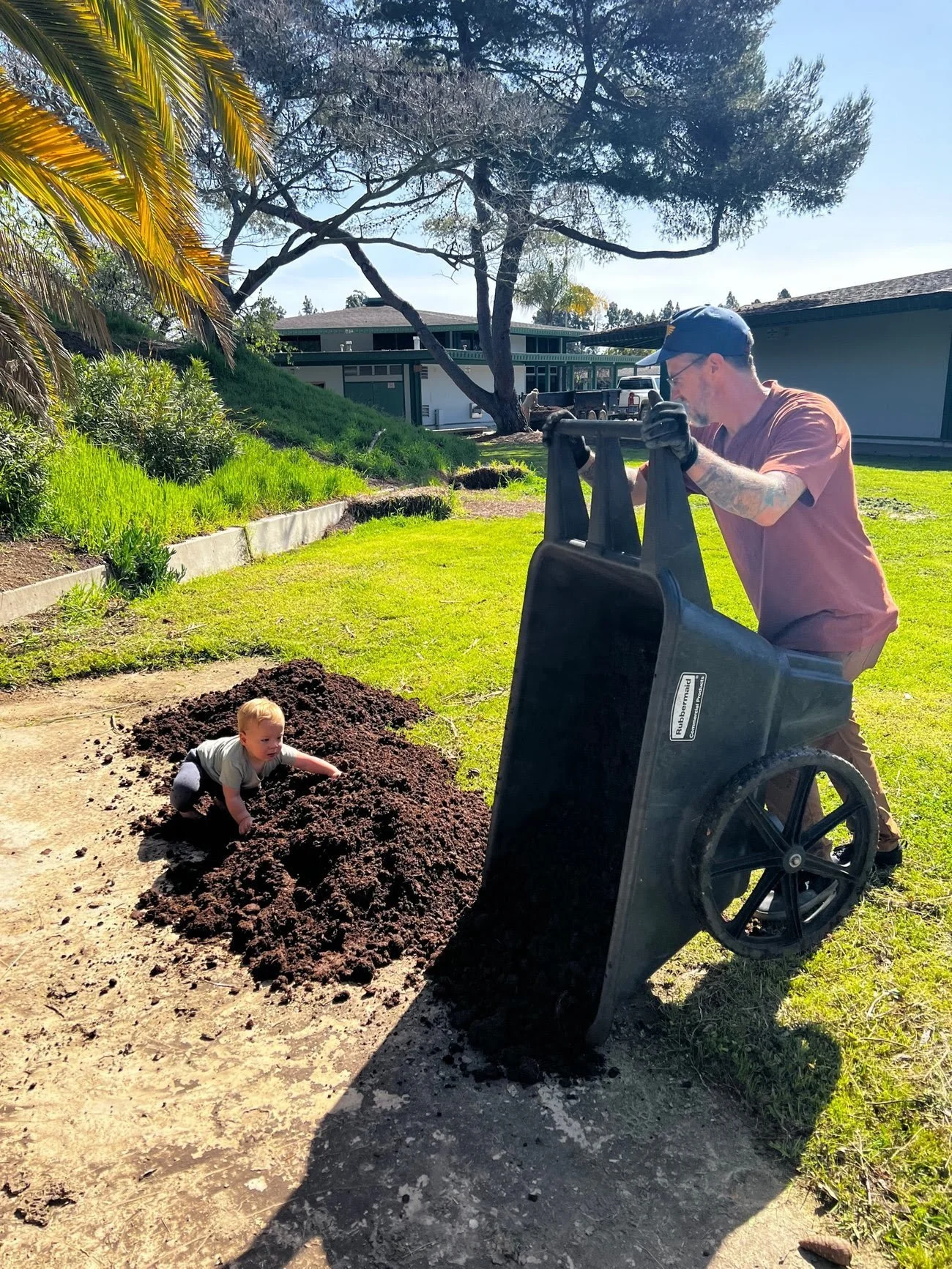 A man pushing a wheelbarrow filled with dark soil while a young child plays in a mound of soil in a backyard on a sunny day.