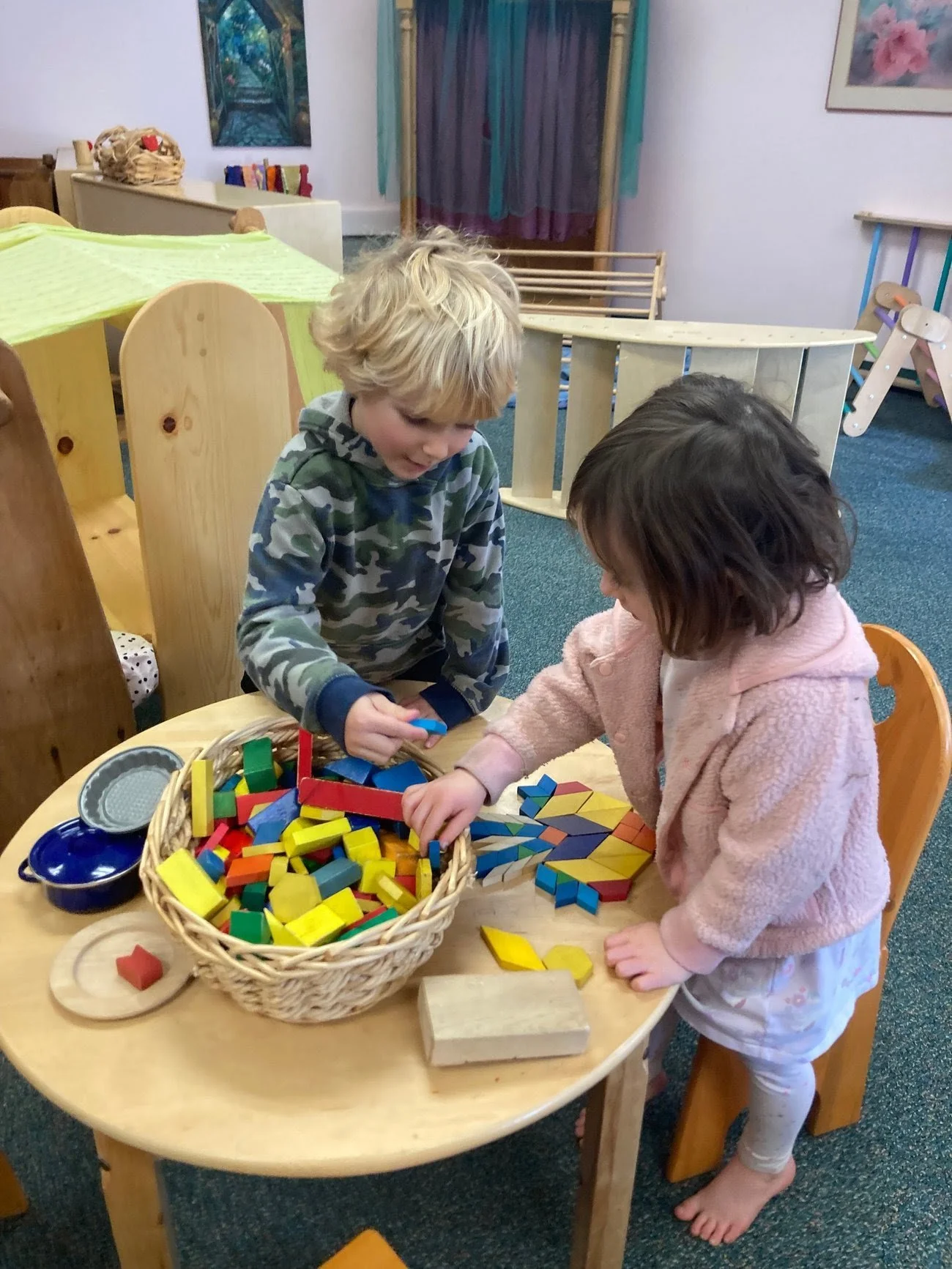 Two children playing with colorful wooden building blocks on a wooden table in a playroom.