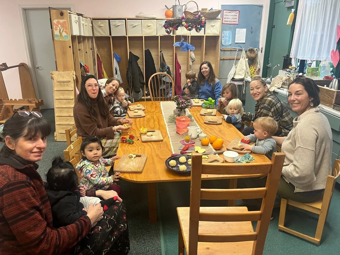 A group of women and children are gathered around a large wooden table in a cozy kitchen or daycare setting. The table has various food items, cups, and oranges on it. The background features open cubbies with hanging coats and bags, and a hanging de
