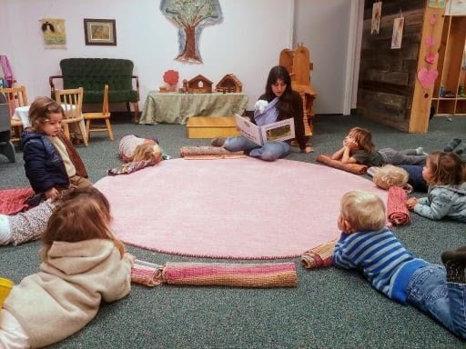 A woman reading a book to children sitting and lying around on a pink rug in a cozy room.
