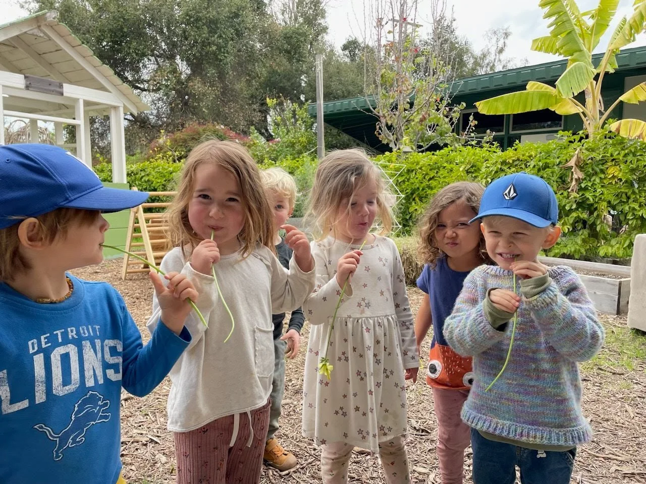 Group of young children, some holding flowers, outdoors in a garden-like setting.