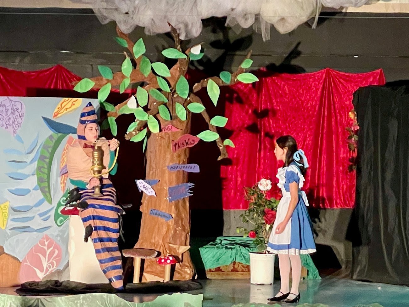 A scene from a play with two young girls in costume, a large paper tree with green leaves, and hand-painted backdrops. One girl in a blue dress with a white apron stands near a potted plant, while the other in a striped outfit with a headpiece sits b