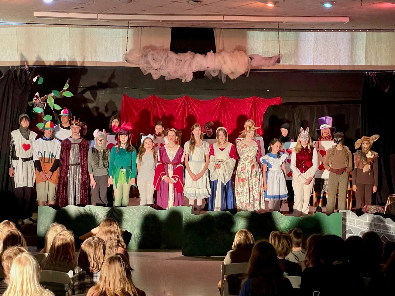 Children's play or theater performance featuring kids dressed in costumes of animals, characters, and storybook figures, standing on stage with an audience watching.