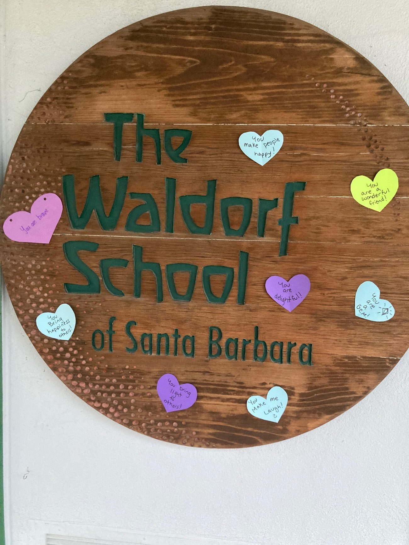 Wooden circular sign with green text reading 'The Waldorf School of Santa Barbara' surrounded by colorful paper hearts with positive messages.