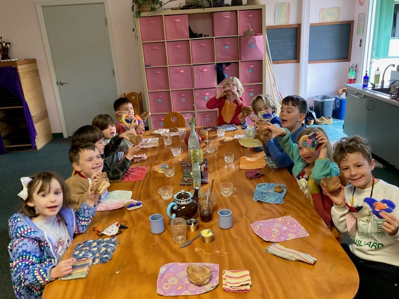 Children sitting around a long table eating baked goods in a classroom setting.