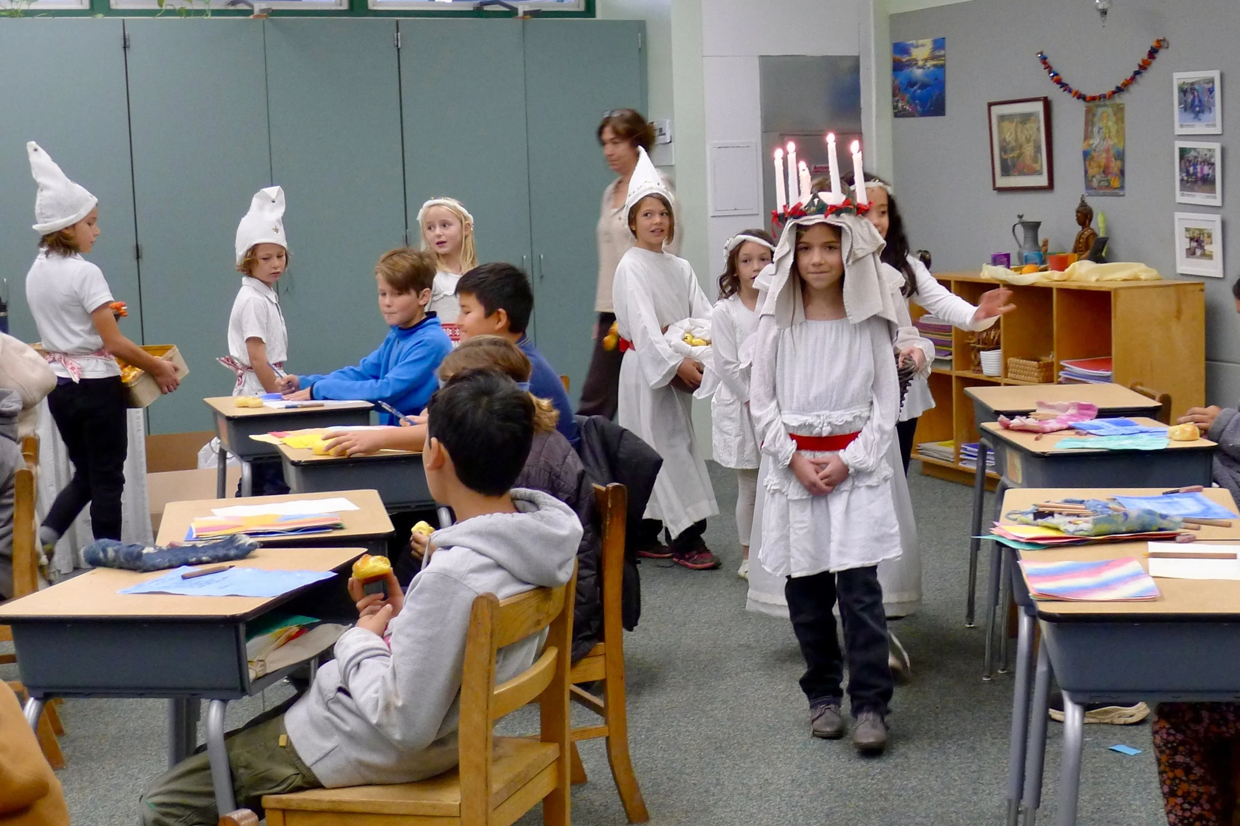Children in a classroom celebrating St. Lucia Day, with some wearing white robes and candles on their heads as part of a traditional Swedish holiday.