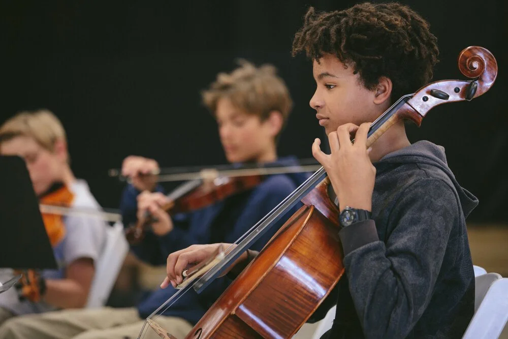 Young boy playing the cello during a music rehearsal with other children in the background.