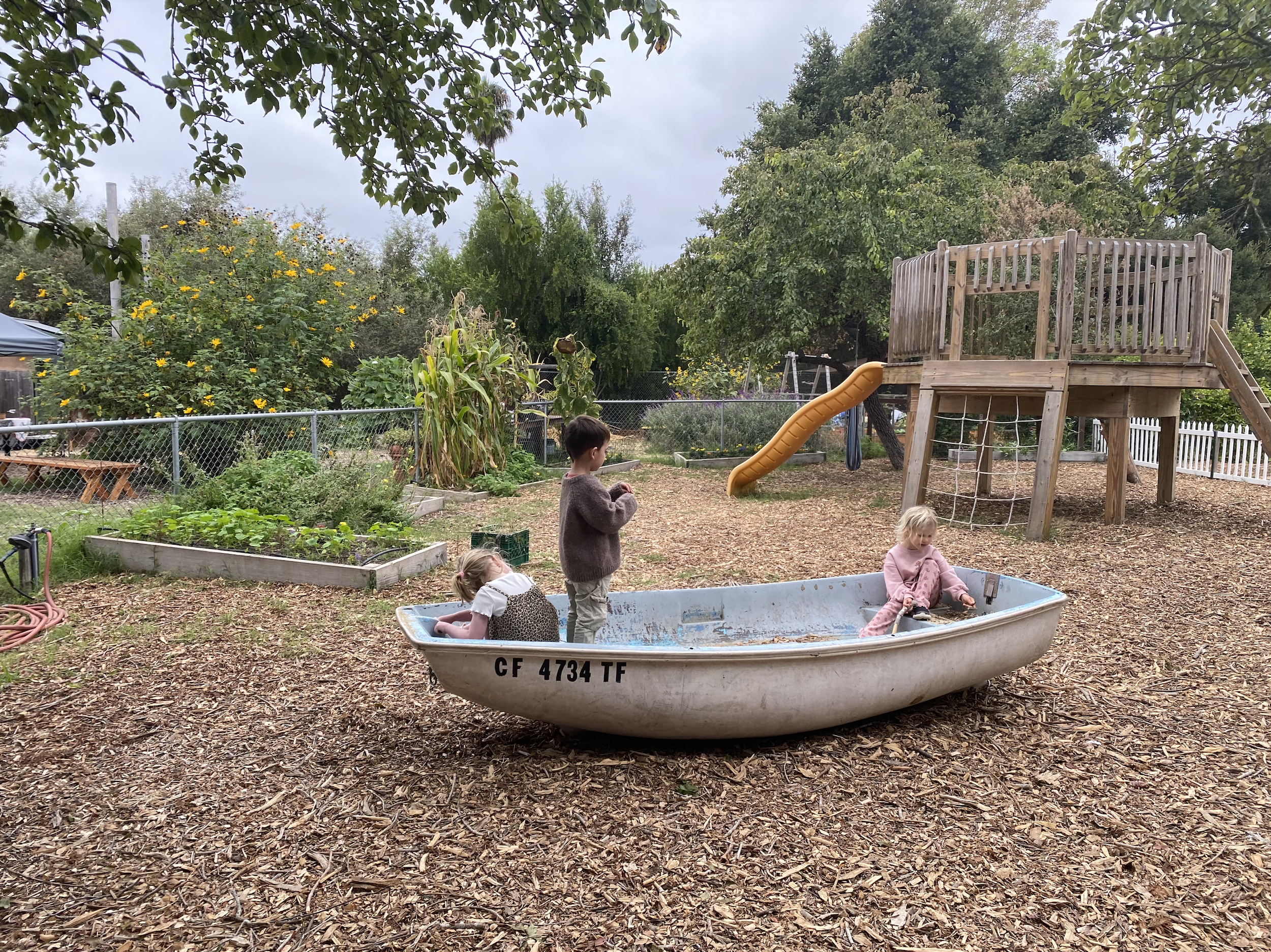 Three children playing in a boat in a backyard playground with a wooden play structure featuring a slide and climbing net, surrounded by trees and garden beds.