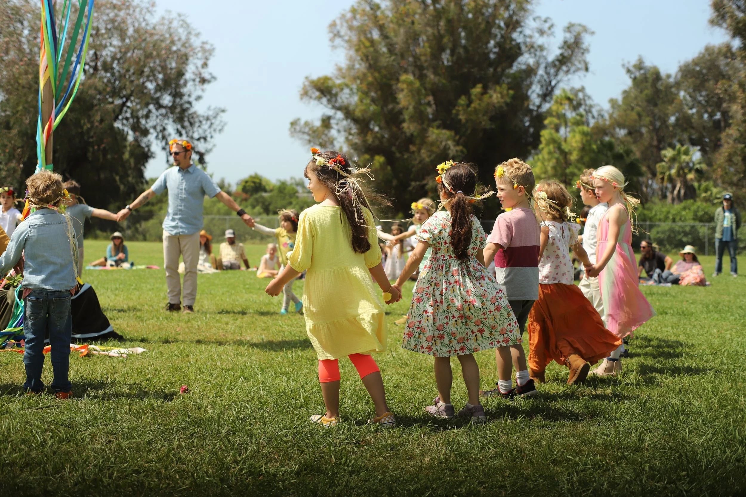 Children dressed in colorful spring clothes holding hands and dancing in a circle outdoors on a sunny day, with adults sitting and standing in the background.