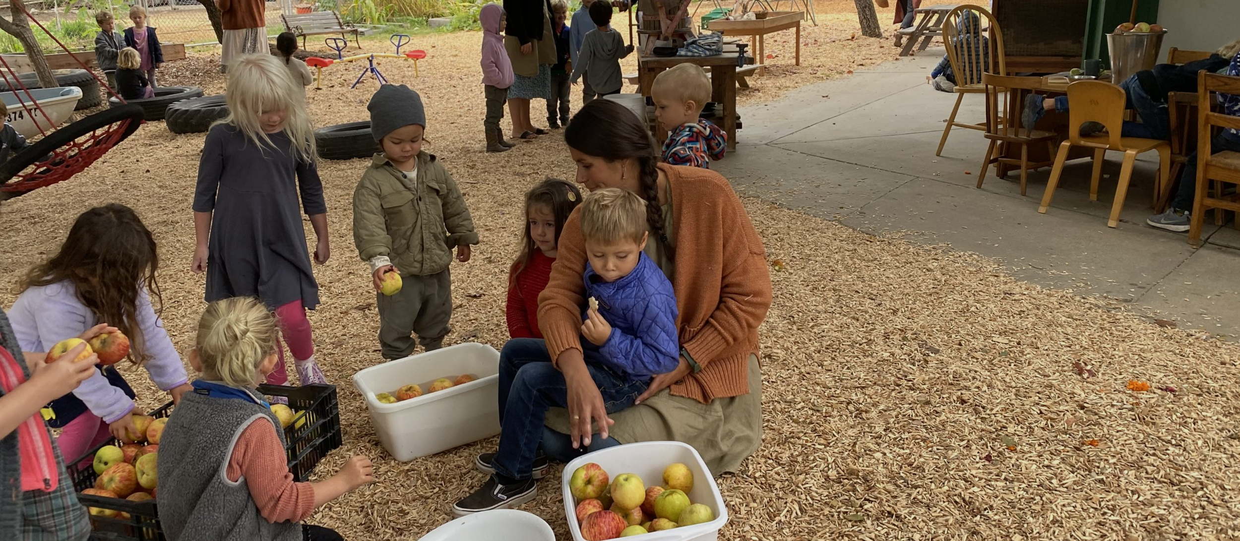 Children and a woman picking apples in an outdoor play area with wood chips, with others playing and sitting at tables in the background.