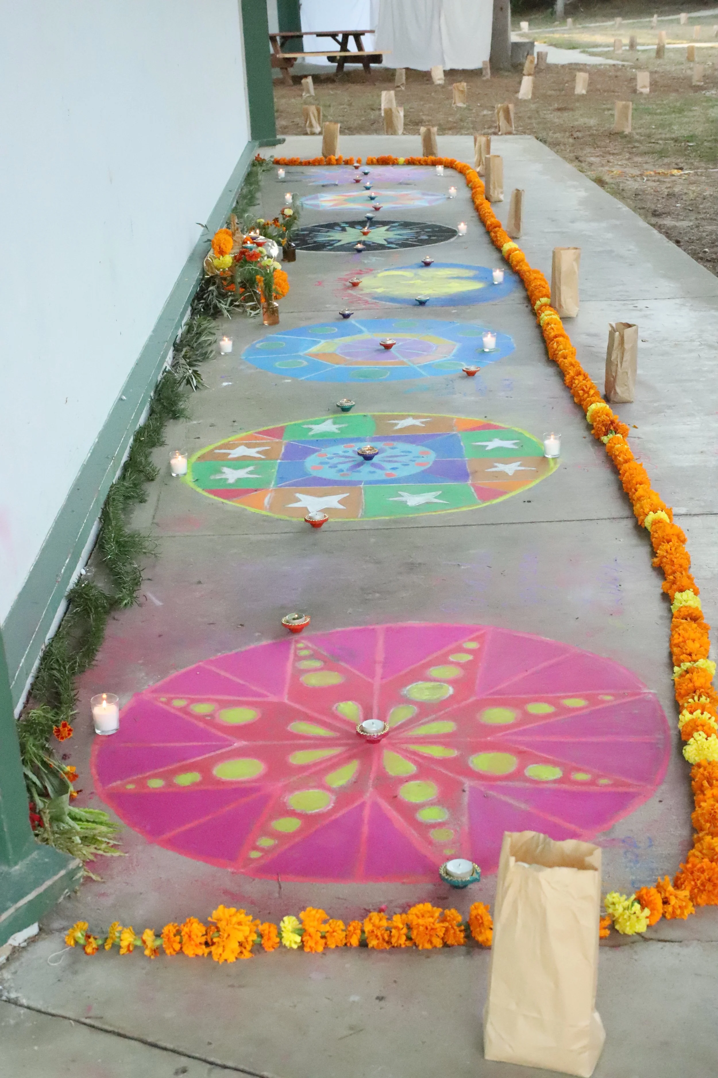 Colorful rangoli patterns on the ground decorated with candles, marigold flower garlands, flowers, and small bags, set up along a pathway outside a building for a celebration or festival.