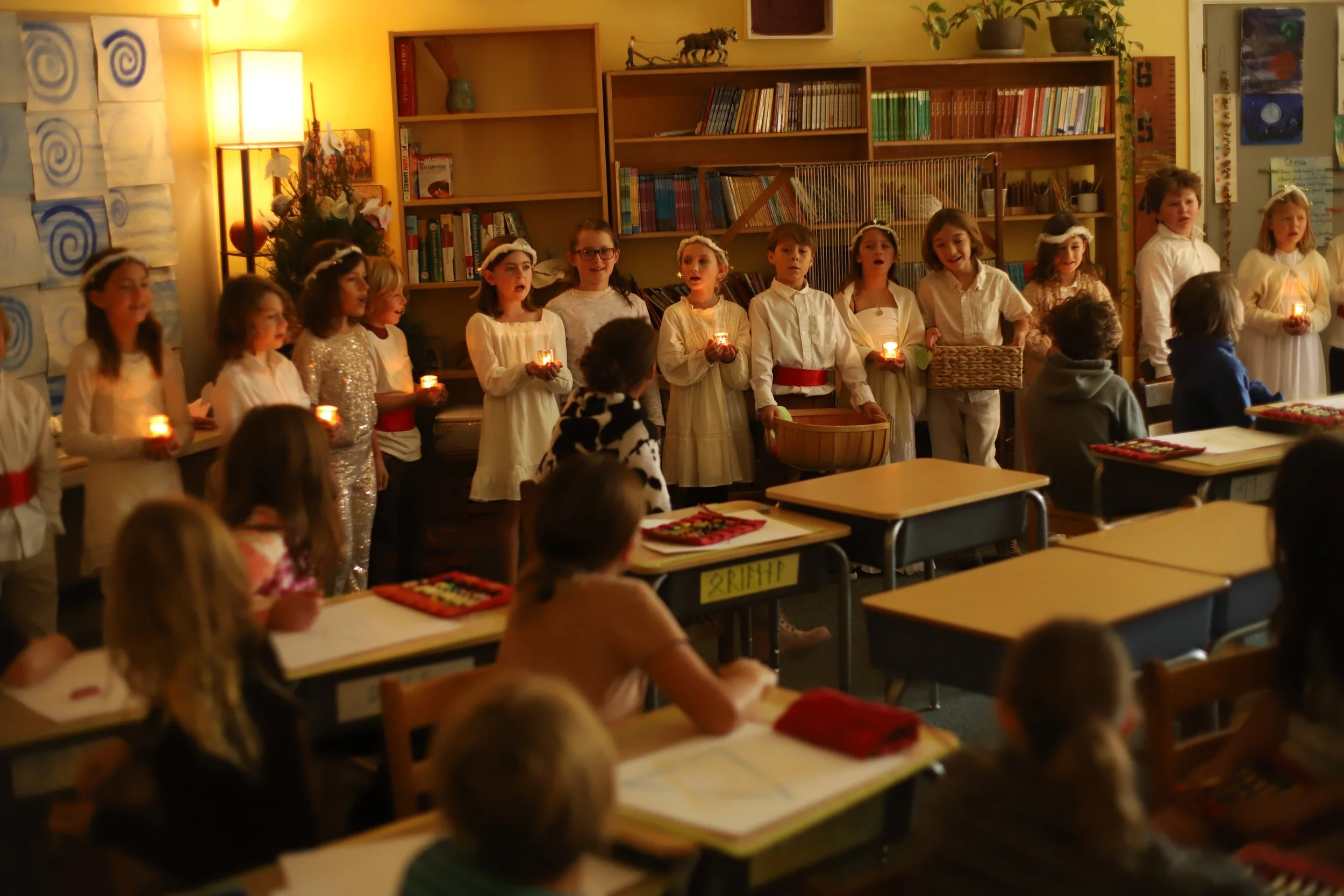 Children dressed in white, some wearing angel costumes, holding candles and performing a Christmas or holiday program in their classroom.