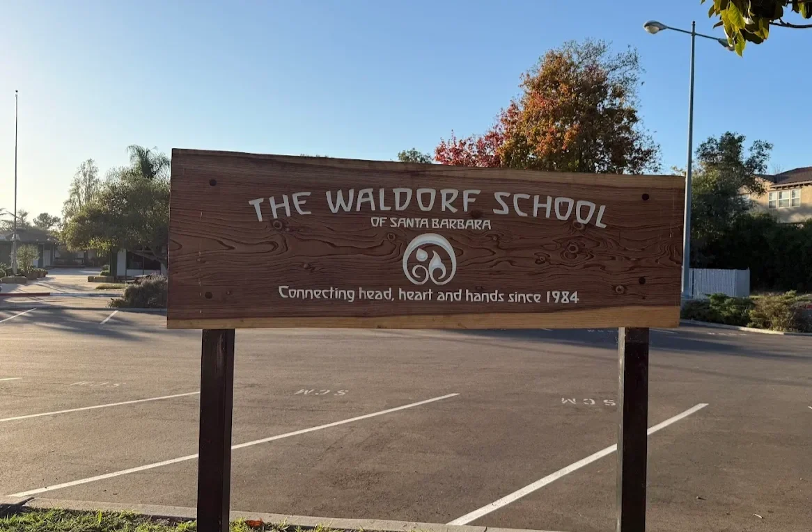 Wooden sign for The Waldorf School of Santa Barbara, with a parking lot in the background and autumn trees.