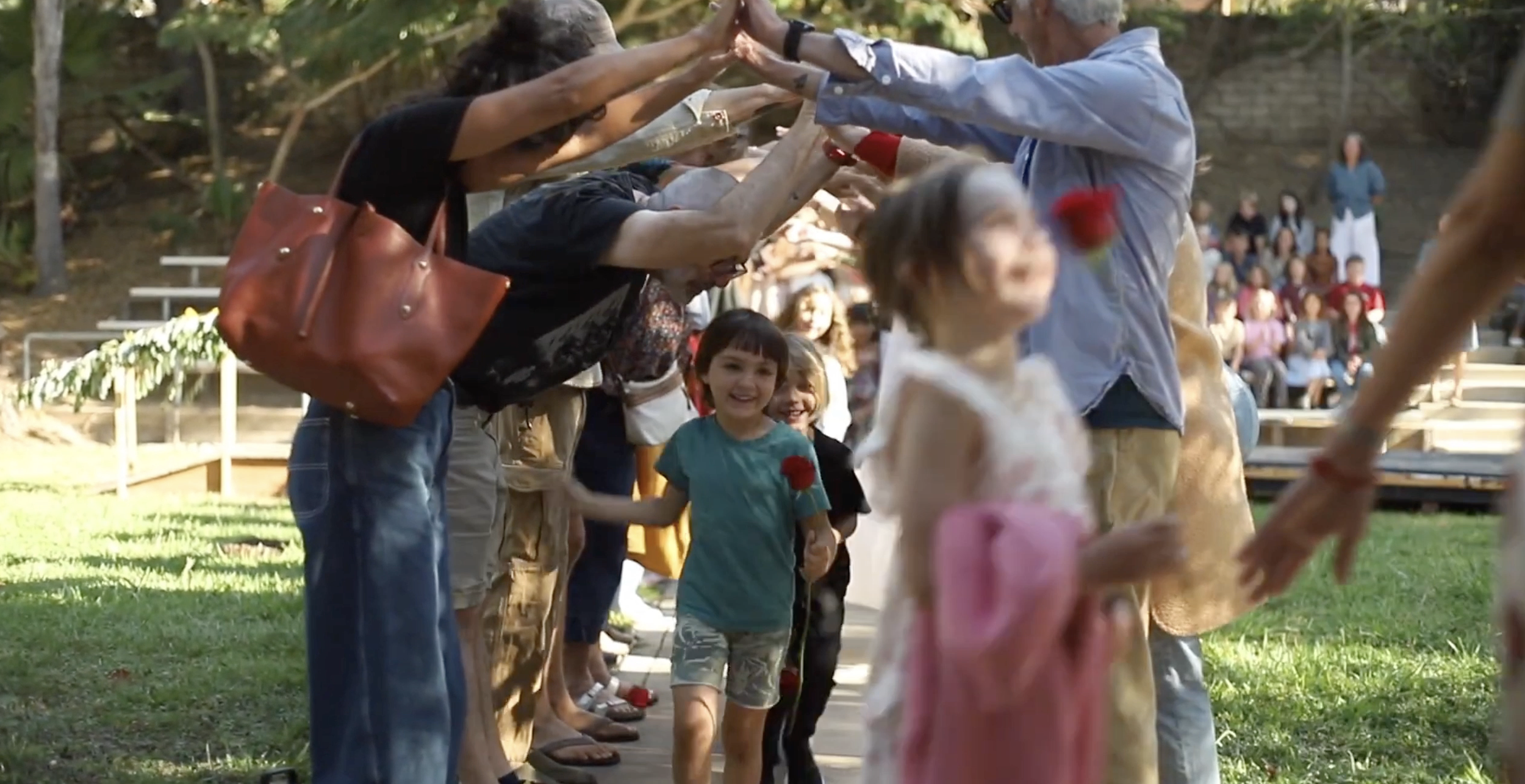 Children holding red flowers and smiling as they walk through a pathway lined with hands raised to form an arch for a celebration or ceremony with an audience seated in the background.
