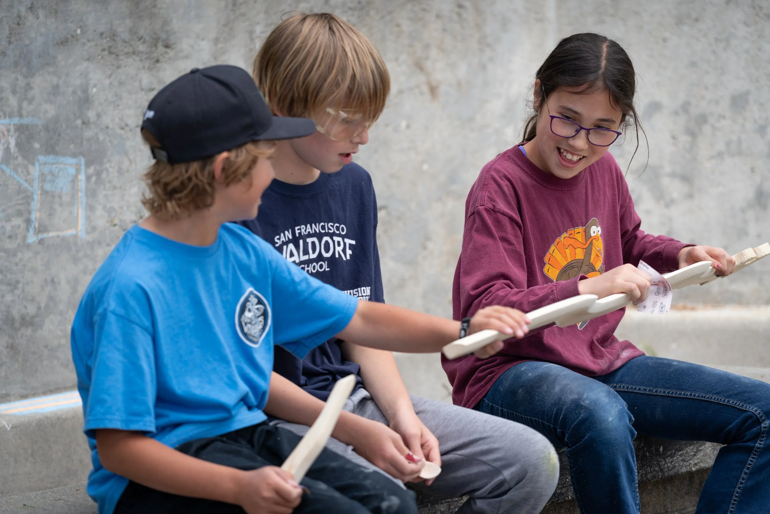 Three kids sitting against a concrete wall, engaged in a game with wooden sticks, smiling and chatting.